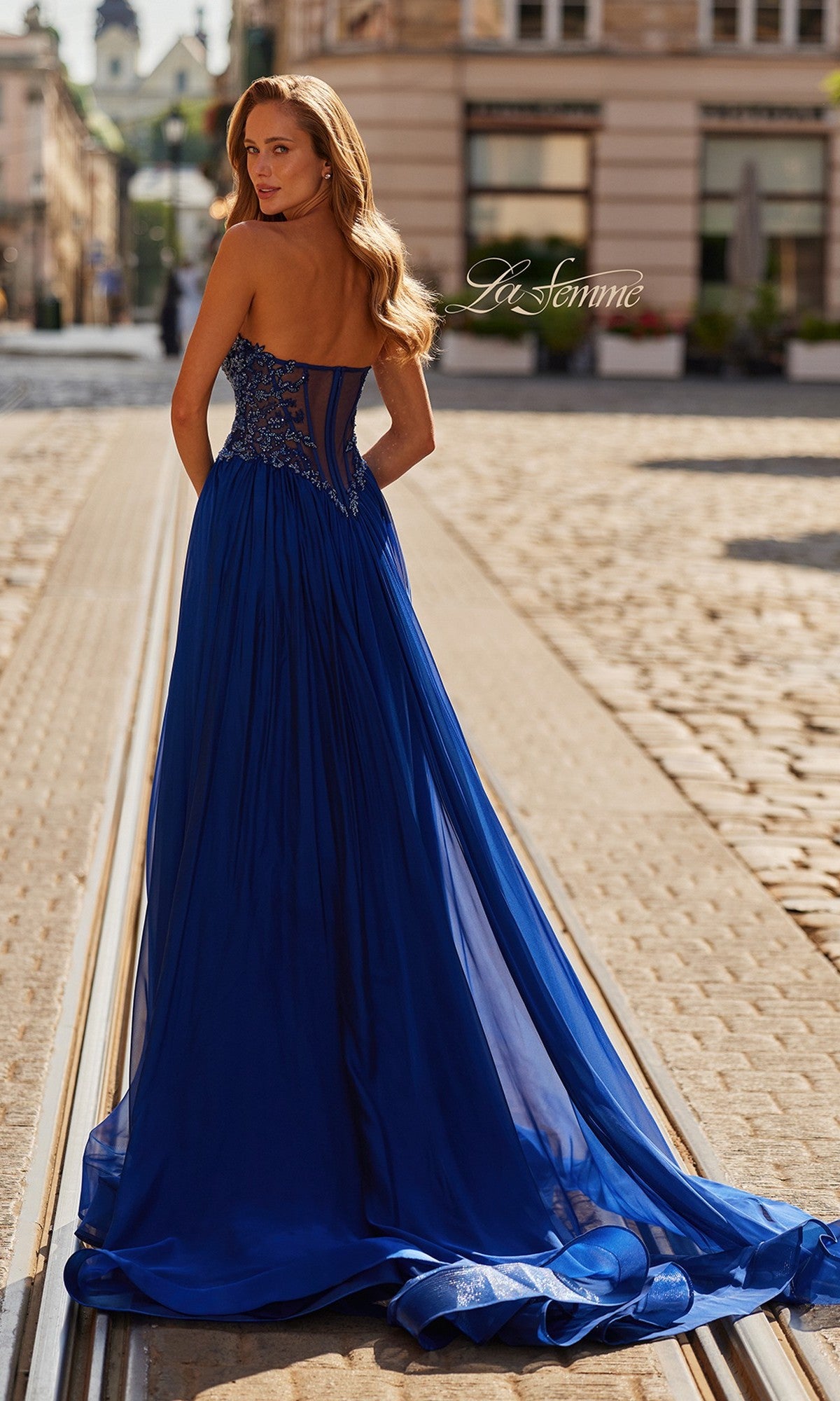 A woman in the La Femme 33552 long prom dress, featuring a strapless chiffon design with a beaded bodice, stands on a cobblestone street and looks over her shoulder.